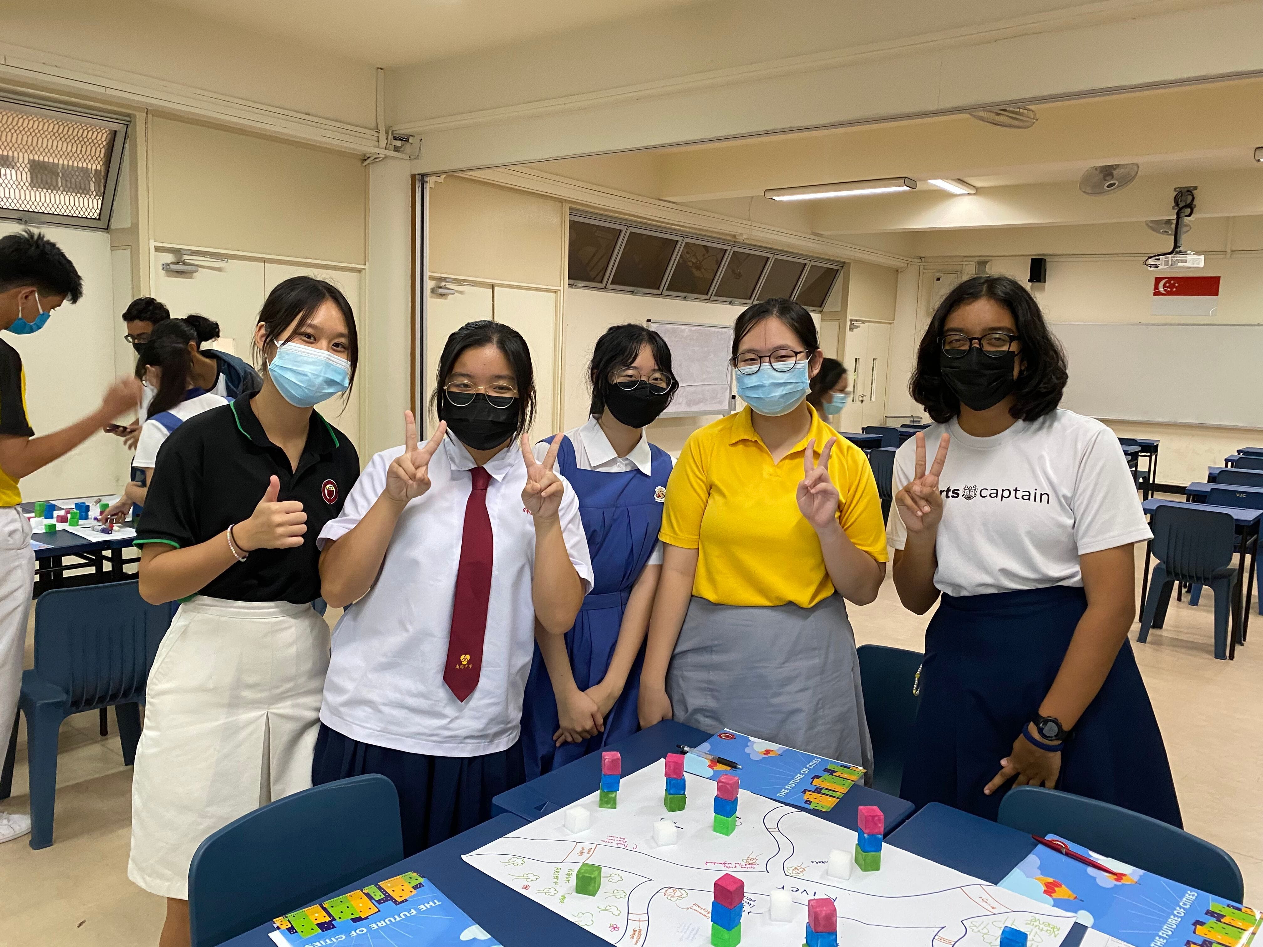 Five masked students in school uniforms pose with peace signs near a table with colorful blocks and a city planning map.